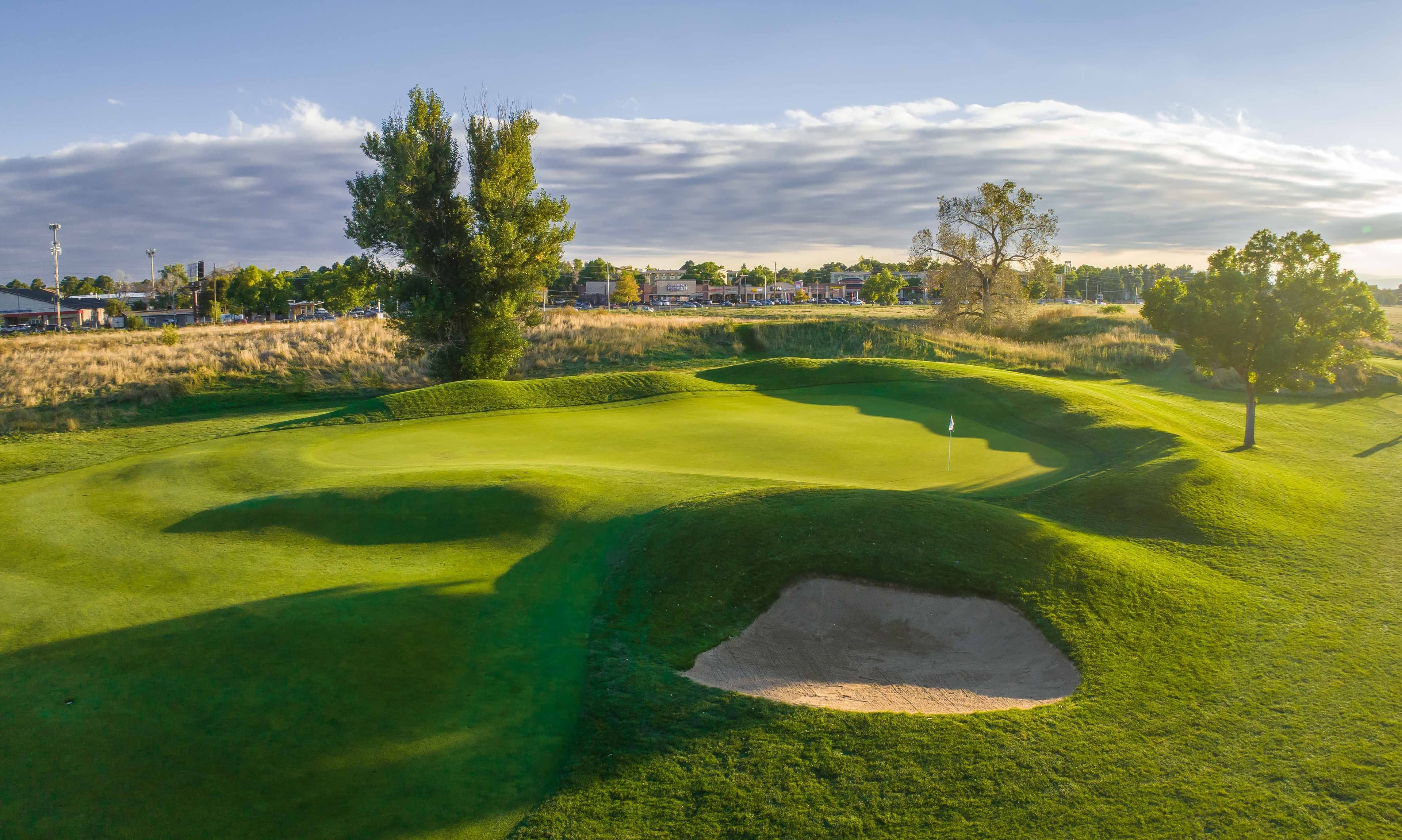 Lush rolling golf green with a sand bunker and white flag at golden hour.